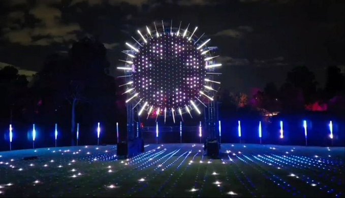 A nighttime outdoor light installation featuring a large circular burst of LED lights above a field of small ground lights and vertical light pillars.