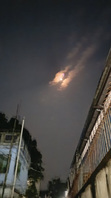 Nighttime photograph of a moon partially obscured by wispy clouds, viewed from a narrow alleyway between buildings.