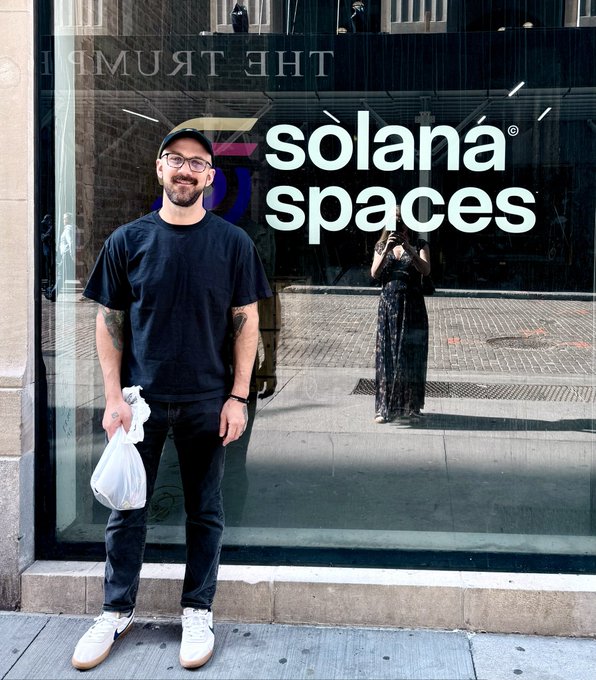 A man in a black t-shirt, jeans, and baseball cap stands smiling on a city sidewalk in front of a storefront window displaying a large "solana spaces" sign, with a woman in a black lace dress visible in the reflection.