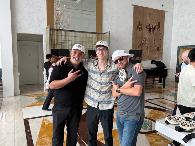 A photo of three smiling men wearing caps posing together in an ornate room with marble floors, with "QUANTUS" branding visible in the background.