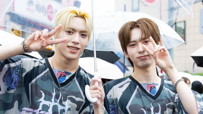 Two young men in matching camo-patterned shirts pose with peace signs while holding clear umbrellas in a rainy outdoor setting.