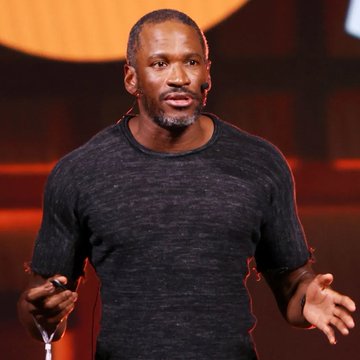 A man with a gray beard wearing a dark t-shirt speaks on stage with an orange-lit background, gesturing with his hands.