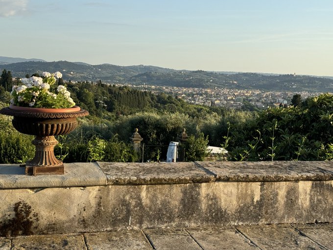 A decorative stone urn filled with white flowers sits on a weathered stone wall overlooking the rolling green hills of Tuscany, with the city of Florence visible in the distance under a soft evening sky.
