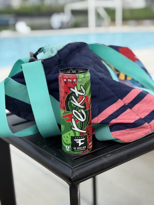 A can of 'NEXT' energy drink in watermelon flavor sitting next to a colorful quilted bag by a swimming pool.