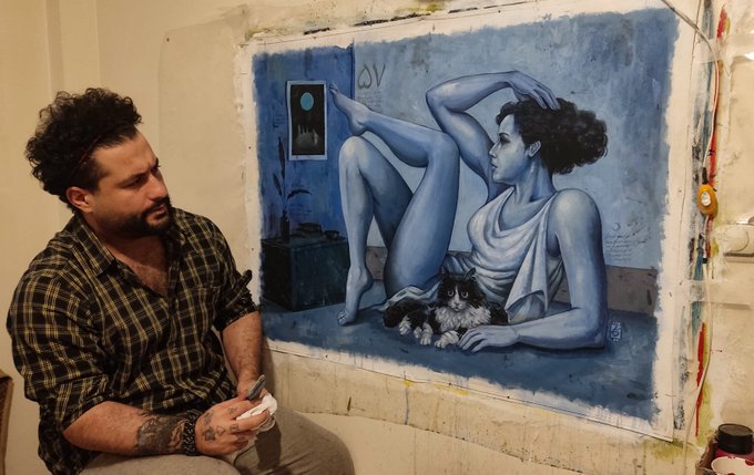 A bearded man sits beside a large blue-toned acrylic painting depicting a reclining figure with a cat, holding a small cup as he contemplates the artwork titled "Nocturne in Blue" (100.70 cm).