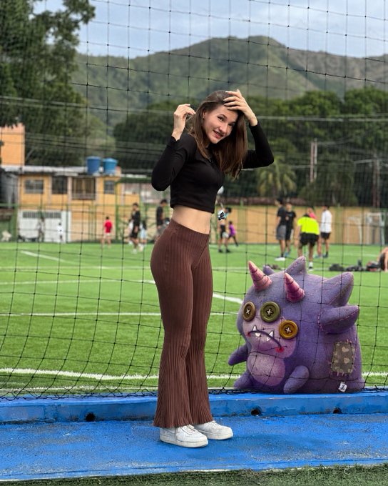 A young woman wearing a black crop top and brown ribbed flared pants posing on a blue track next to a purple plush monster toy, with a sports field and mountains in the background.