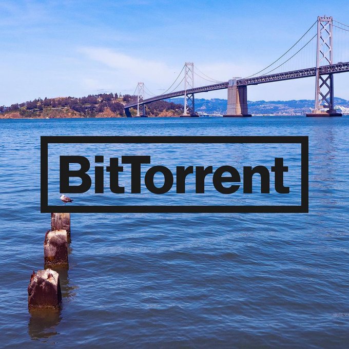 The image shows a scenic view of the San Francisco Bay with the Bay Bridge spanning across the water, featuring wooden posts emerging from the blue water in the foreground, with a seagull perched on one post. The word "BitTorrent" is prominently displayed in bold black text within a rectangular border overlaid on the water, visually reinforcing the post's theme of BitTorrent's expanding reach and influence in decentralized data sharing and storage technology.