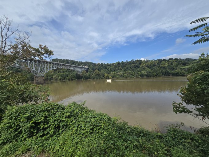 A scenic landscape photograph showing a steel arch bridge spanning a wide brown river, with a small white boat visible on the water, surrounded by lush green forested hills under a partly cloudy blue sky.