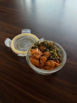 An overhead view of a clear plastic container holding a meal with tofu and leafy greens, resting on a dark wooden table.
