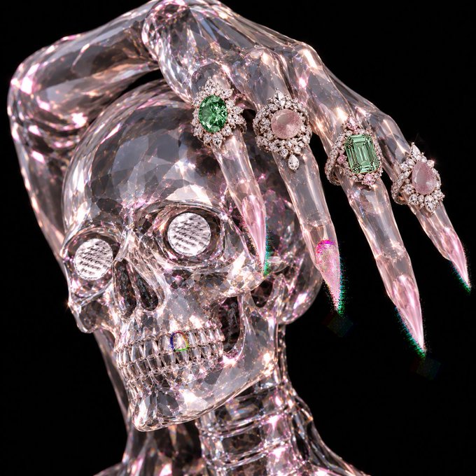 A close-up of a clear crystal skull sculpture featuring a faceted hand resting on top, adorned with jeweled rings and gemstones set into the eye sockets.