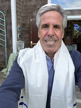 A smiling man with gray hair wearing a navy blue shirt and a white towel or scarf draped around his neck poses for a selfie outdoors near a building.