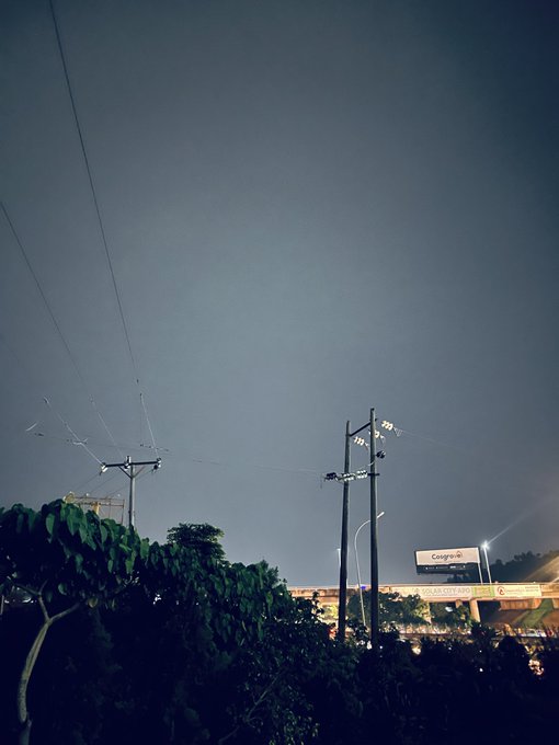 An evening or dusk photo of an urban skyline with electrical transmission towers and power lines in the foreground, silhouetted trees below, and a bridge or elevated road with a billboard reading 'Cogere' in the distance under a gray sky.