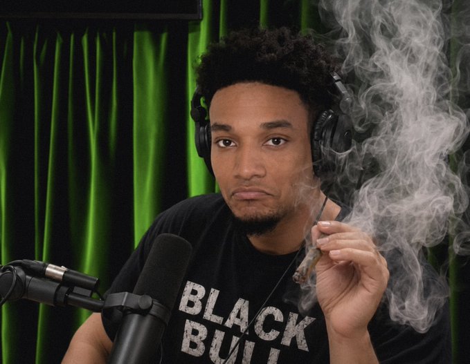 A young man with curly hair wearing a 'BLACK BUILT' t-shirt and headphones, smoking a cigar with visible smoke, seated in front of a microphone against a green curtain backdrop.