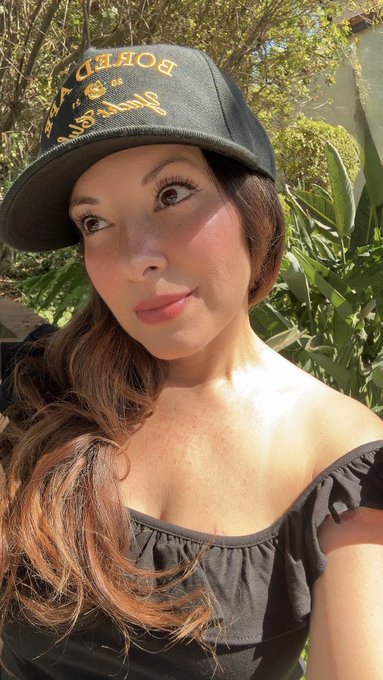 A woman wearing a black cap with gold lettering that appears to read 'BORED' and a black off-the-shoulder top, posing outdoors near green foliage.