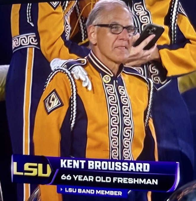 A television broadcast shows Kent Broussard, identified as a '66 YEAR OLD FRESHMAN' and 'LSU BAND MEMBER,' wearing a yellow and purple LSU marching band uniform while holding up a smartphone.
