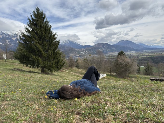 The image features a woman lying down on grass, possibly enjoying her time outdoors or taking a break from work. She has her legs crossed and appears relaxed as she looks out at the surrounding area. The background includes mountains, suggesting that they are likely in a mountainous region. There is also a tree visible near the woman, adding to the natural setting of the scene. The image does not contain any text or other objects, so it focuses on the main subject - the woman lying down and enjoying her time outdoors.