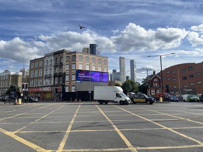 A street-level photograph of a London intersection (likely Shoreditch area) featuring a digital billboard, mixed architecture with modern skyscrapers in the background, a bird flying in a partly cloudy sky, and a white delivery van driving through yellow box junction markings on the road.
