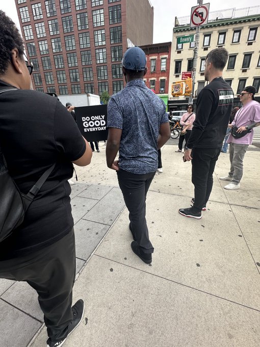 Several people stand on a city sidewalk, including a man holding a black banner that reads 'DO ONLY GOOD EVERYDAY' with buildings in the background.