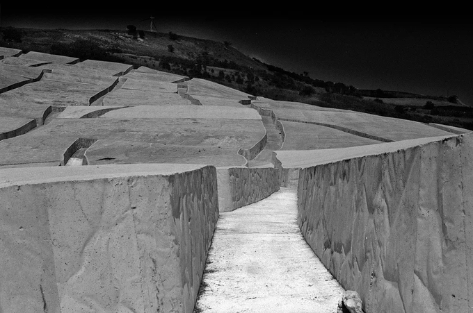 A dramatic black-and-white photograph captures the monumental concrete fissures of Il Grande Cretto, Land Art artist Alberto Burri's memorial sculpture in Sicily, carved into the ruins of the destroyed town of Gibellina. The narrow pathway between towering white concrete slabs evokes the haunting atmosphere that inspired this digitally minted tribute on the Manifold platform.