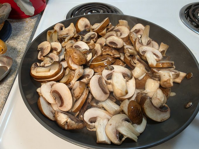 A black skillet on a white stove is filled with sliced cremini and button mushrooms being sautéed.