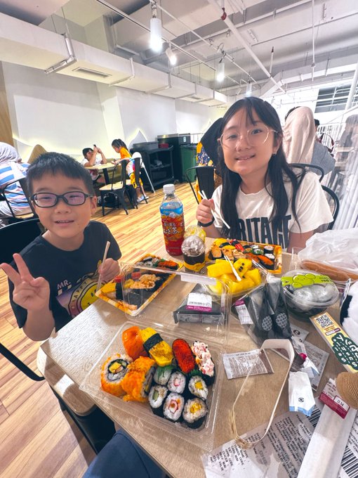 A young boy and girl smile at the camera while posing with peace signs behind a table full of sushi bento boxes and snacks in what appears to be a cafeteria or food court.