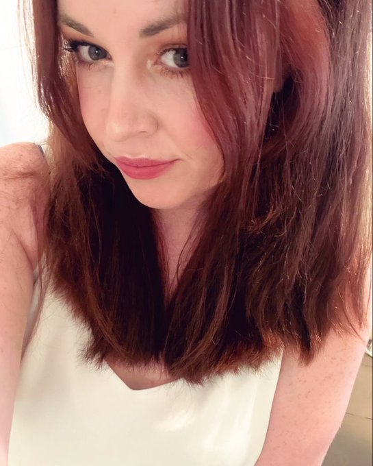 A close-up portrait of a young woman with long reddish-brown hair wearing a white tank top, looking directly at the camera.