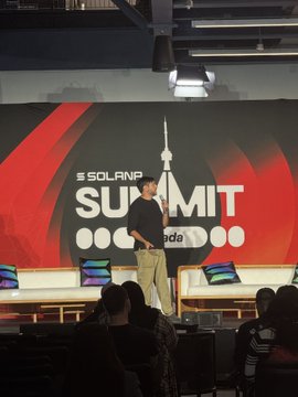 A presenter stands on a stage at the 'SOLANA SUMMIT' event, with an audience seated in the foreground.