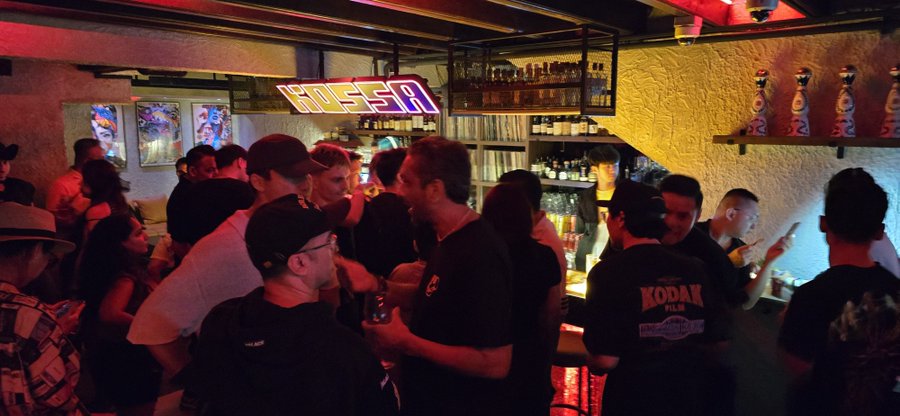A crowded bar scene at night under red lighting, with a neon 'HOESR' sign above the bar and a patron wearing a 'KODIAK' t-shirt.