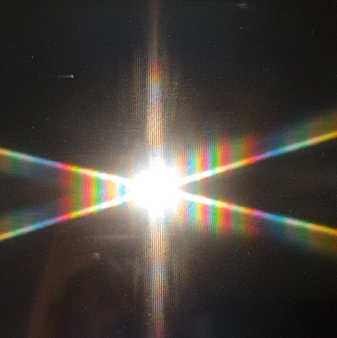 A photograph capturing a bright point of light creating a colorful starburst or diffraction spike effect against a dark background.