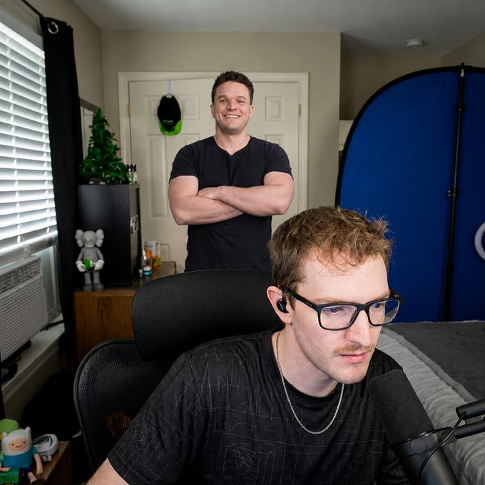 Two men are pictured in a bedroom setup that appears to be a streaming or content creation space, with one seated in front of a microphone wearing glasses and earbuds while the other stands behind him with arms crossed, smiling at the camera. A laptop displays the Stake logo, fitting the caption's request for viewers to share their Stake usernames.