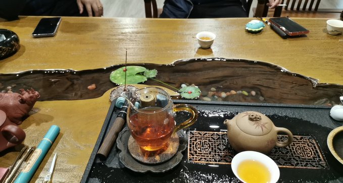 The image shows a wooden table with various objects placed on top of it, including cups, bowls, and other items. There are two people sitting at the table, one person on each side. The table also has a teapot or tea kettle, which is likely used for making tea. A cell phone can be seen on the table as well. The post that accompanies this image provides information about the objects in the photo and their significance.