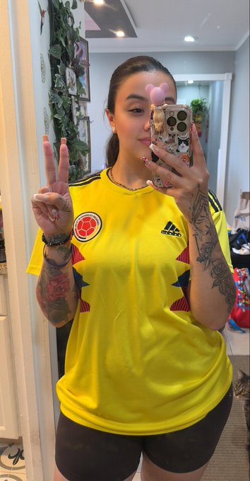 A woman with arm tattoos takes a mirror selfie wearing a yellow Colombia national football jersey with the Adidas logo and federation crest, flashing a peace sign while holding a pink phone case. The caption celebrates a 161-day selfie streak awaiting MON's all-time high, with the Colombian kit matching the post's prideful "Colombia, sí, sí Caribe" chant.