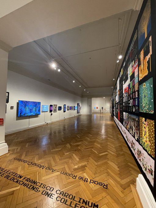 A spacious art gallery interior with wooden floors and walls lined with various artworks; floor text reads "Read the Room - Alma And Wood Collections, FRANCISCO CAROLINUM COLLECTION, TATSIS: ONE BLACKCHART".