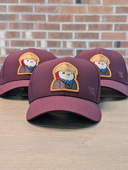 Three maroon/burgundy baseball caps are displayed on a wooden surface, each featuring an embroidered patch of a character wearing a yellow hat.