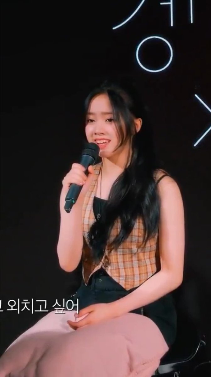 A young woman with long dark hair, wearing a plaid vest over a black top, smiles while holding a microphone during what appears to be a fan event or talk show. Korean text at the bottom reads "의치코 싶어" (partial), and the hashtags #UNIS #유니스 #ELISIA #엘리시아 identify her as Elisia from the K-pop group UNIS.