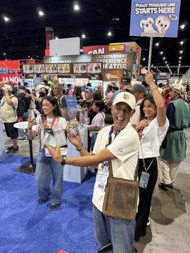 A woman in a white t-shirt and cap holds up merchandise while smiling at a crowded convention floor, with visible signage reading "Plushy Pendants are STARTS HERE" and "FAN EXHIBITOR EXPERIENCE SEARCH."