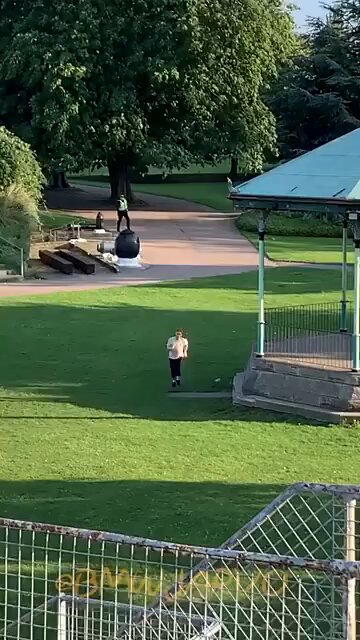 The image shows a person walking through a park with a green roofed gazebo nearby. The scene includes a bench located near the gazebo, as well as some trees in the background. There are also two other people present in the park, one of whom is carrying a backpack. A fence can be seen in the foreground, and there's a post with text on it that reads "Be smart, USE #Monero! They are getting mad just because you use it, imagine if you HODL $XMR".