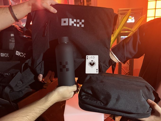 A pair of hands display an OKX-branded merchandise set featuring a black bag, a matching water bottle, and an Ace of Spades playing card, all set against a red-lit venue. The image ties directly to the post's celebration of receiving the "spade ace" gift package from the OKX event.