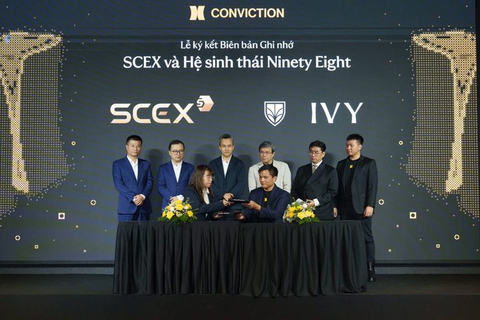 A group of people standing behind a table on a stage during a signing ceremony (Lễ ký kết Biên bản Ghi nhớ) between SCEX and the IVY ecosystem, under the CONVICTION logo.