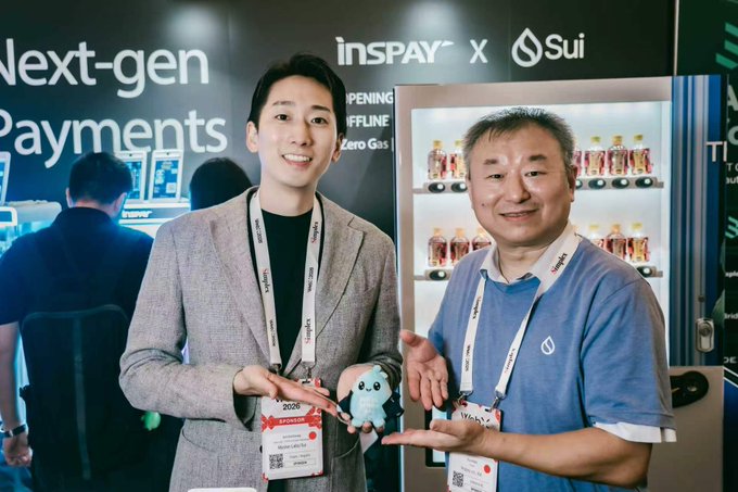 Two men pose at a trade show booth for 'insPAY X Sui' promoting 'Next-gen Payments,' with one holding a small blue plush mascot.