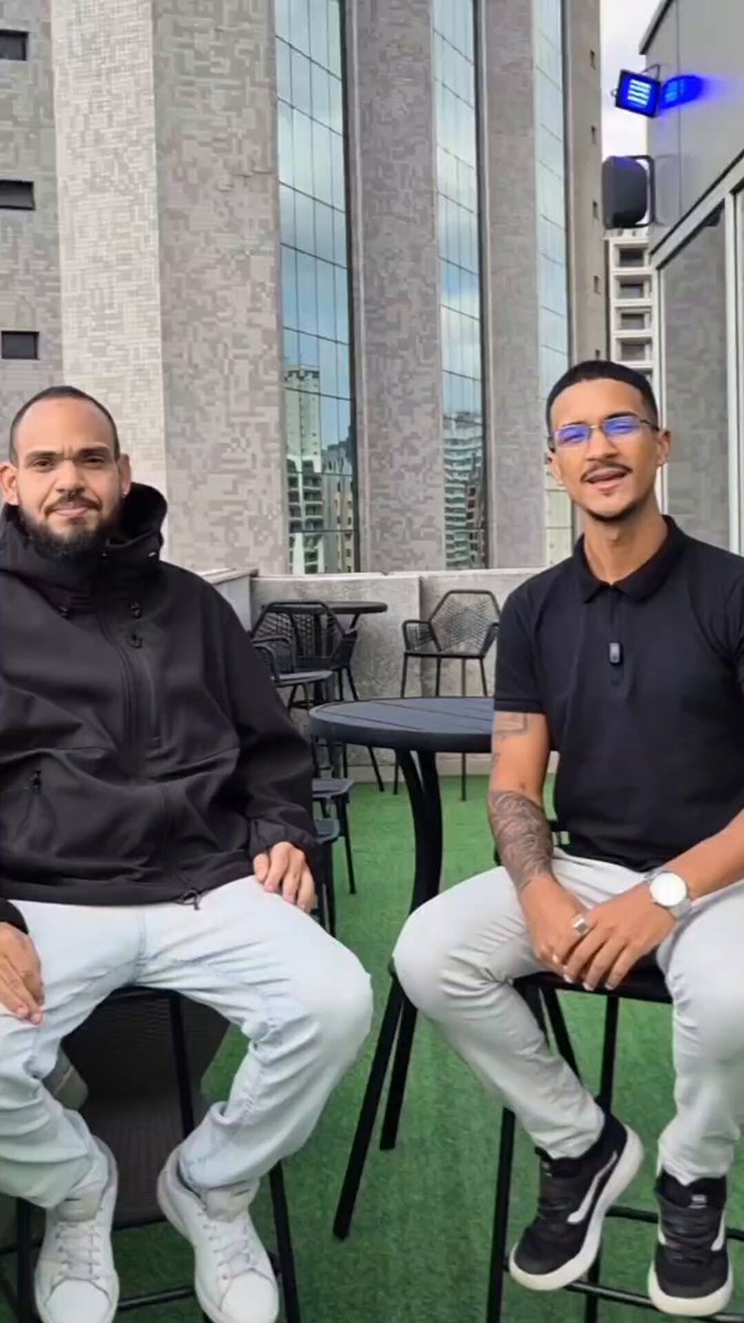The image features two men sitting at a table, both wearing black clothing. They are positioned next to each other with their legs crossed over one another. In the background, there's a building that appears to be made of concrete or stone, possibly an office building. The photo also contains text that reads "Stellar Perfect Storm!!!". This suggests that the image might have been taken at a location where the two men are attending a meeting or event related to the post "Stellar Perfect Storm!!!".