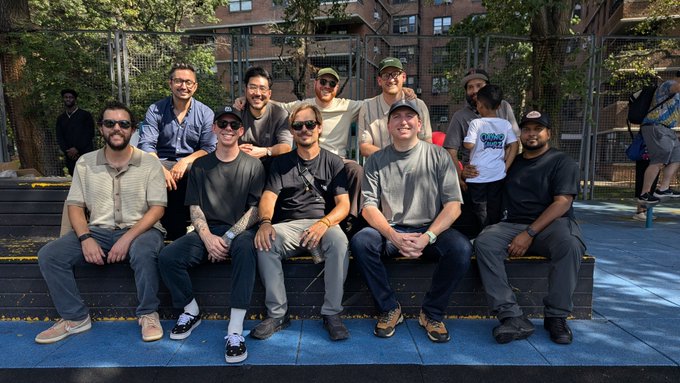 A group of about ten men posing together on bleachers at an outdoor basketball court, dressed casually in t-shirts, hats, and sneakers.
