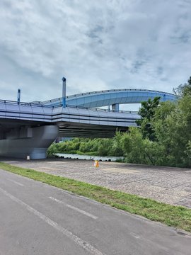 An outdoor view of a bridge over a river with green vegetation along the banks, taken from a roadside path on a cloudy day.