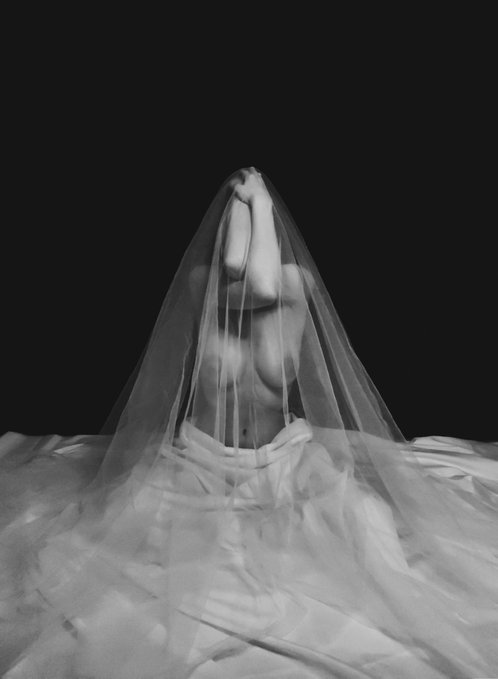 A black and white artistic photograph shows a person partially draped in a sheer veil against a dark background, with a satin fabric at their waist.