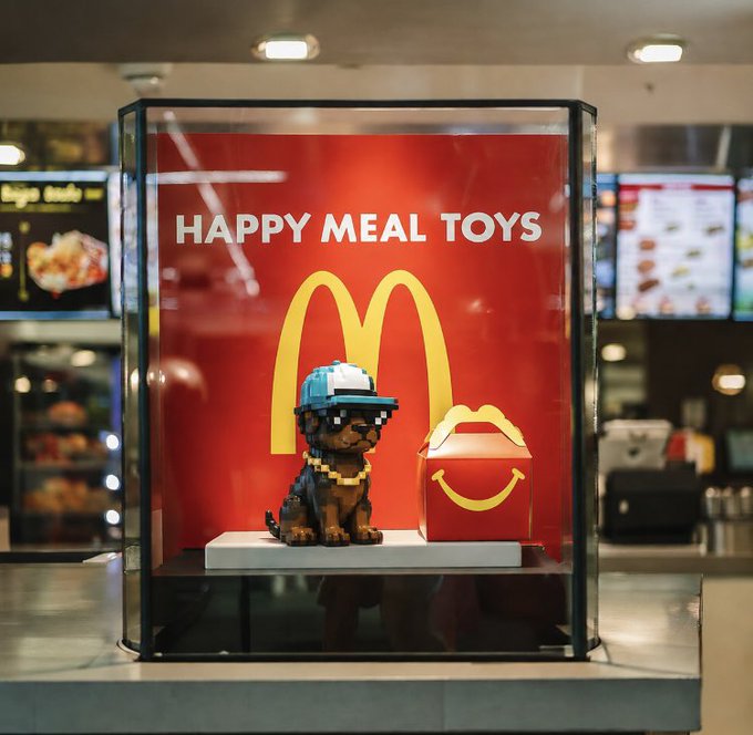 The image features a display case at a McDonald's restaurant, showcasing a small dog wearing sunglasses and a hat, sitting next to a red box with a yellow logo that says "McDonald's Happy Meal Toys". The display case is located near the bottom of the frame, while the background shows other displays in the restaurant. The post about accepting Dogecoin D o O nly G ood E veryday suggests that the image might be related to a promotional campaign or event at McDonald's, possibly encouraging customers to use cryptocurrency for their purchases.

The dog and the Happy Meal toys are positioned close together on a glass shelf in the display case, drawing attention to the items inside. The overall scene conveys a sense of fun and excitement associated with the McDonald's Happy Meal toy offerings.