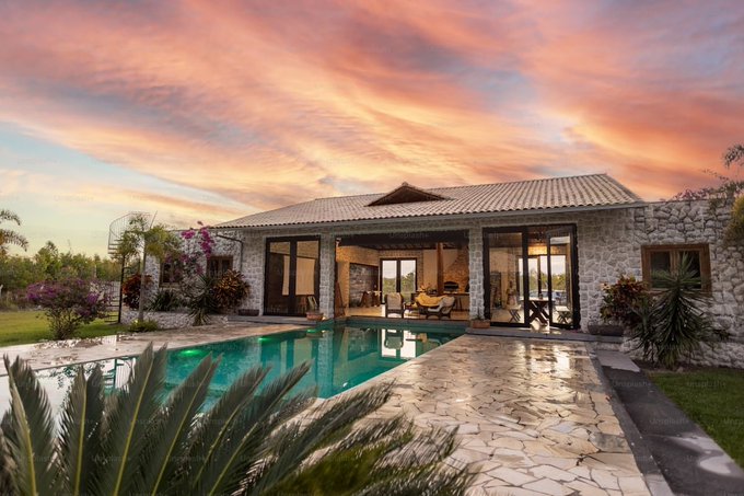 The image shows a large house with a pool outside of it. The sky has pink clouds, creating an interesting backdrop for the scene. There are also palm trees visible near the house, adding to the tropical atmosphere. The photo captures the essence of the property, showcasing its unique features and inviting ambiance.