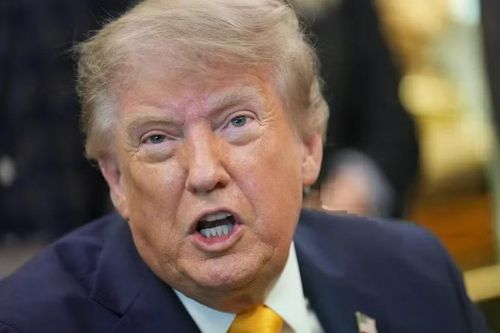 A close-up photo of Donald Trump speaking, wearing a dark suit with a gold tie, captured mid-sentence.