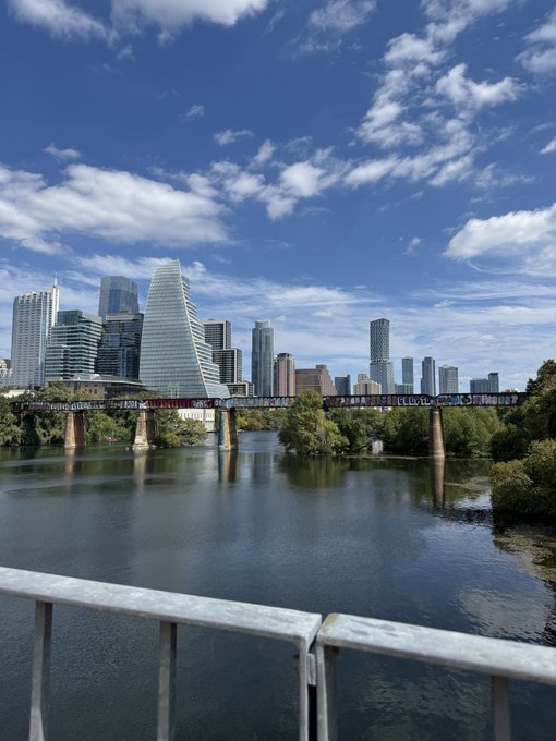 A scenic view of a city skyline featuring modern skyscrapers behind a railway bridge spanning a calm river, with a blue sky dotted with clouds above.