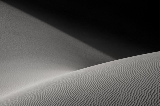 A black and white close-up photograph showing the textured, rippled surface of sand dunes with contrasting light and shadow.