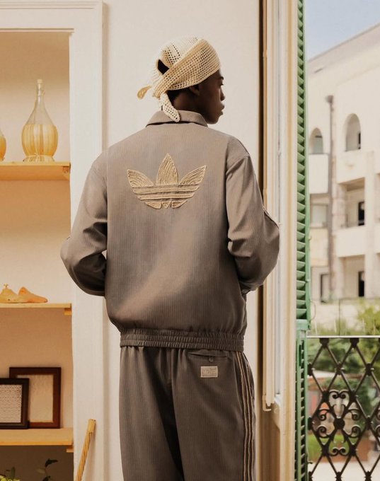 A person wearing a gray Adidas Originals tracksuit and a crocheted headwrap stands by an open doorway looking out at buildings.